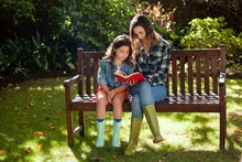 Woman Reading Book To Daughter While Sitting On Wooden Bench
