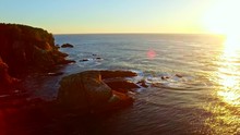 Sweeping Reveal Of A Sunset At Pacific Ocean, Starting With Rocky Cliffs With Mountains In Background, Rotating Around A Small Rocky Pillar Ending With Wide, Cinematic, Colorful, Golden, Coastal Ocean