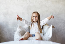 Portrait Of Charming Happy Little Girl With Bare Feet And Long Straight Hair Sitting On White Armchair, Showing Thumbs Up With Both Hands As Sign Of Approving And Liking Good Idea. Body Language