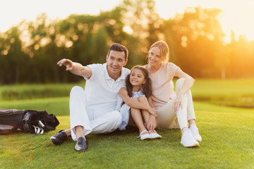 The family sits on the grass on the golf course, the man points to the side, and the wife and daughter look there