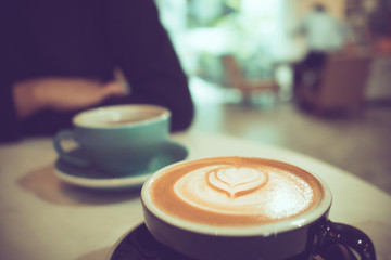 two cups of hot coffee at cafe, one with heart shape latte art, man with black long sleeve shirt as background, selective focus, vintage tone