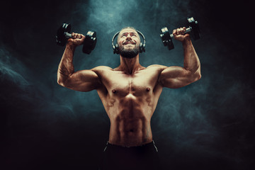 athletic man training muscles with dumbbells in studio on dark background with smoke. Strong bodybuilder with six pack, perfect abs, shoulders, biceps, triceps and chest posing with headphones.