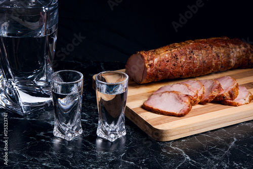 Zdjęcie XXL Butelki i kieliszków z wódką z plastrami wędzonego mięsa na drewnianą deską do krojenia.