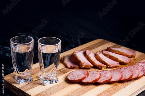 Zdjęcie XXL Kieliszki do wódki z wódką, plastry wędzonego mięsa i wędzonej kiełbasy na desce do krojenia.