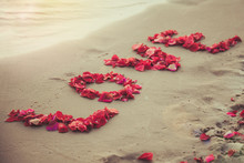 Word Love Made Of Red Rose Petals. Symbol Of Love. Screen Saver For Love Story. Background For Text About Wedding And Wedding Trips. Flower Petals In The Shape Of The Word Love On The Sand Beach.