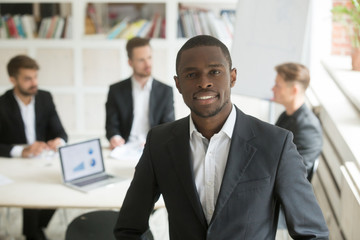  Headshot portrait of handsome young smiling african american businessman looking at camera. Blurry team of male businessmen on background, Friendly male executive, CEO, corporate worker or manager.
