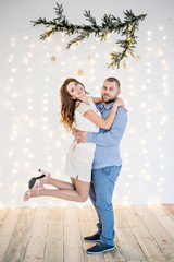 A loving couple have fun on the background of a white wall with garlands of bright lights, a branch of a Christmas tree hangs from above. Lovers are happy dancing at the party waiting for the new year