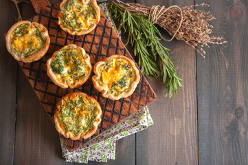 Savory mini quiches (tarts) on a wooden board. Flaky dough pies. Fresh rosemary and dry thyme on a wooden background.