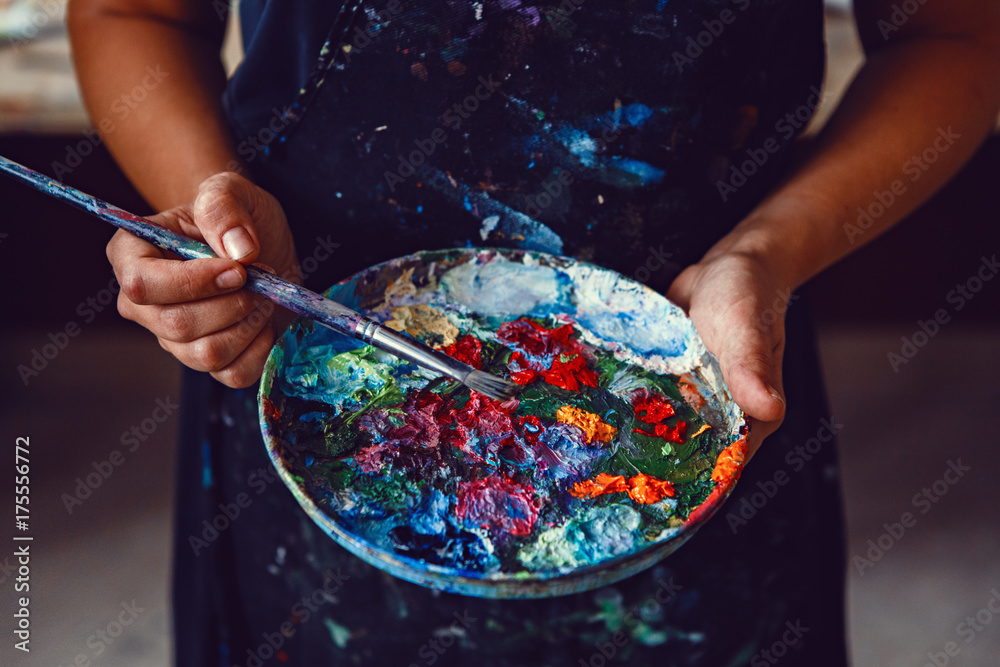 Hands of female artist holding messy dirty palette  with different paints and paintbrush in art studio. Lifestyle and hobby concept - obrazy, fototapety, plakaty 