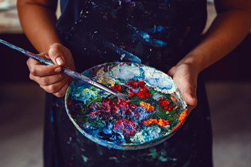hands of female artist holding messy dirty palette with different paints and paintbrush in art studi