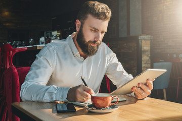 Wall Mural - Young bearded businessman sits in cafe, home at table, uses tablet computer and writes in notebook. Man is working, student is studying. Online education, marketing, training. E-learning, e-commerce.