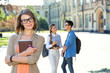 © A Stock Studio - Young female student holds textbook. Woman standing in the background of a group of students on the university campus. Student life.