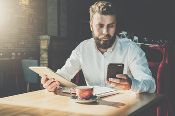 Wall Mural - Young serious bearded businessman sits in cafe at table, holds digital tablet, uses smartphone.There is notebook on table,cup of coffee.Online education,marketing,training.E-learning.Instagram filter.