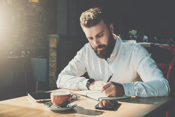 Young bearded businessman sits in cafe at table and writes in notebook.On table tablet computer, smartphone.Man is working, planning.Online education,marketing.E-learning, e-commerce.Instagram filter.