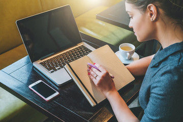 Sticker - Side view. Young businesswoman sitting at table in cafe in front of laptop, computer and making notes in notebook, diary. Girl working, student studying. Online marketing, education, e-learning.