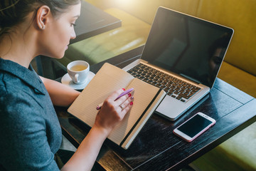 Sticker - Side view. Young woman sitting at table in cafe in front of laptop, computer and making notes in notebook, diary. Businesswoman working, student studying. Online marketing, education, e-learning.