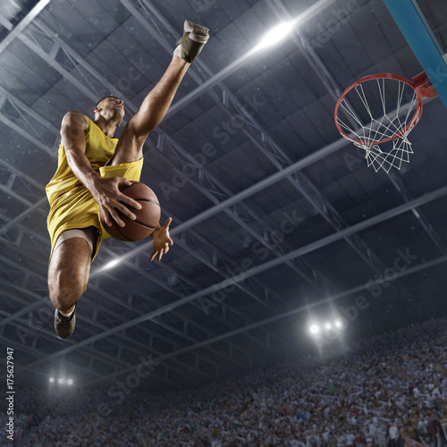 Plakat Gracz koszykówki robi slam dunk na dużej profesjonalnej arenie. Gracz leci w powietrzu z piłką. Gracz nosi ubrania niemarkowe. Widok z dołu.