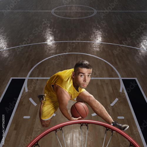 Plakat Gracz koszykówki robi slam dunk na dużej profesjonalnej arenie. Gracz leci w powietrzu z piłką. Gracz nosi ubrania niemarkowe. Widok z góry.
