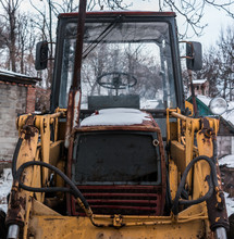 Old Rusty Tractor With Large Wheels Stands In A Snowy Yard. Winter Time