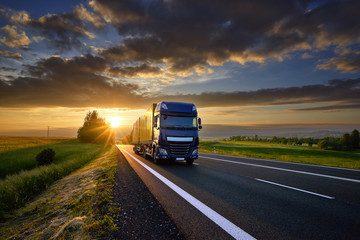Wall Mural - blue truck driving on the asphalt road in rural landscape at sunset between dark clouds