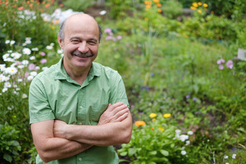 An elderly man with a mustache and a bald spot in a green T-shirt is standing among flowers in the summer garden, arms crossed and smiling.