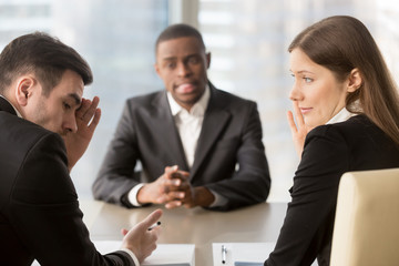 Skeptical female and male caucasian employers covering face with hand and whispering while discussing african american candidate. Disappointed hr managers secretly consulting on failed job interview