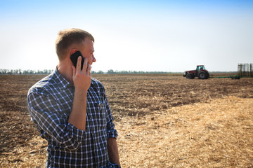 Young man with phone in a field and a tractor on a background.