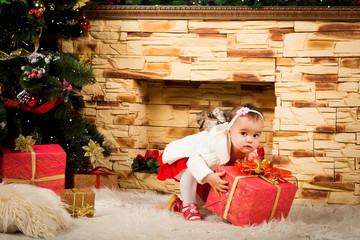 Wall Mural - Little blonde girl with a cute crown in a white smart dress in New Year room near the fireplace and the beautiful large Christmas tree decked out with gold, red and silver toys