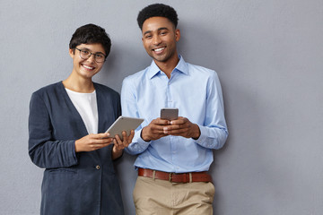 Female and male enterpreneurs have meeting at office, consult internet using electronic devices, smile gladfully, demonstrate positive emotions. Young managers create business project together