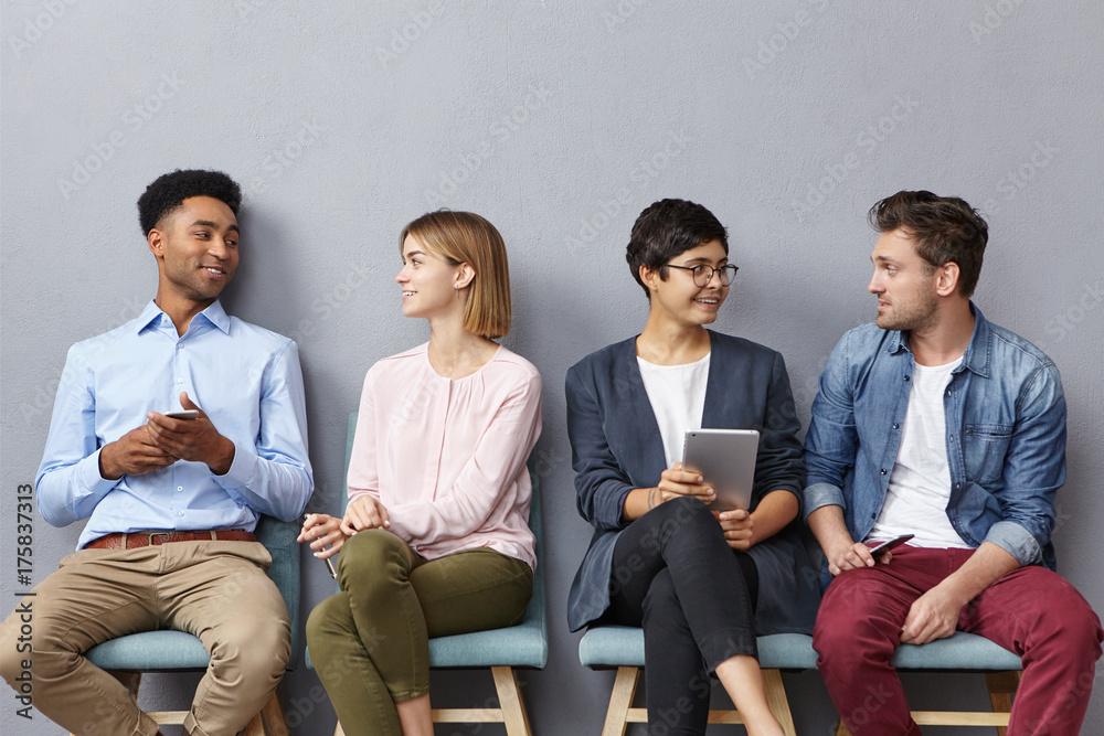 Horizontal portrait of people sit in queue, have pleasant conversation with each other, share ideas and life experience, isolated over grey concrete wall. Diverse group in row, speak and hold gadgets