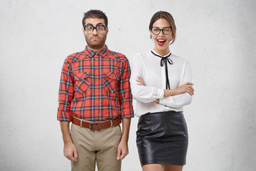 Wall Mural - Best friends stand next to each other: unshaven puzzled man in glasses and checkered shirt and beautiful woman winks with eye joyfully, being glad to meet former schoolmate, remember school years