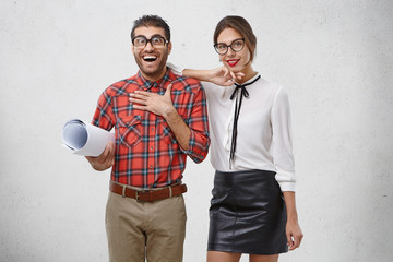 Wall Mural - Excited unshaven young man doesn`t believe his eyes, keeps hand on chest, holds rolled up paper or drawing which man prepared with beautiful female. Team work, cooperation and surpriment concept