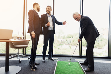 Wall Mural - A respectable elderly man is playing golf in his office. Next to him are his workers. They look like the boss is playing