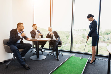 Wall Mural - Three men sit at a table and watch a woman in a business suit playing mini golf in the office