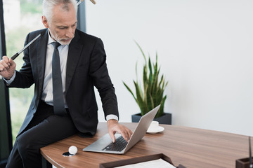 Wall Mural - An elderly respectable man posing in his office with a golf club. He sits on the desktop behind the laptop