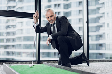 Wall Mural - A respectable elderly man playing a mini golf in the office. He smiles and looks at the miniature golf course