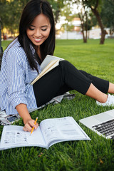 Sticker - Happy asian female student in striped shirt writing to notebook, while studying in park, outdoor