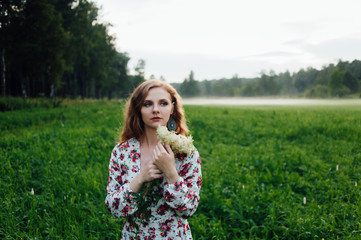 A beautiful girl in a colorful dress stands in the evening on a meadow against a background of fog.