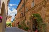 Fototapeta Uliczki - Beautiful narrow street with sunlight and flowers in the small magical and old village of Pienza, Val D'Orcia Tuscany, Italy.