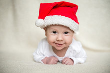 Little Baby In Red Cap Of Santa Claus Celebrates Christmas. Christmas Photo Of Infant In Red Cap. New Year's Holidays And Christmas Tree.