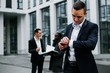 © pantovich - Young businessman in the foreground looks at the clock on the background of colleagues on a city street.
