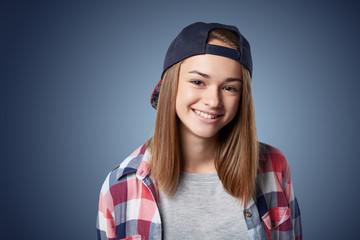 Wall Mural - Closeup portrait of smiling teen girl wearing checkered shirt and baseball cap, over grey background