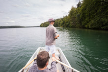 A Man Is Rowing A Rowboat While His Friend Is Fly Fishing From The Boat On A  Lake.