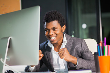 Businessman at office, success gesture, goal reached, happy man. Hardworking male worker. Hipster afro american man at light office with computer monitor. Satisfied with job done