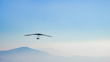 Hang Glider Soaring The Thermal Updrafts Suspended On A Harness Below The Wing, Misty Mountain Peaks And Blue Sky