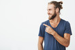 Profile of cheerful handsome man with fashionable hairstyle and beard smiling brightfully and pointing at free space for advertisement.