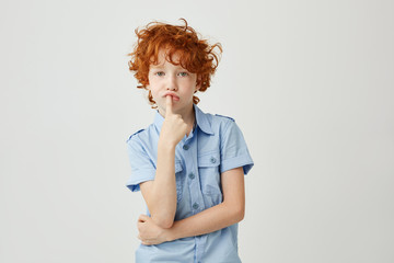 Wall Mural - Portrait of funny little child with ginger wavy hair and freckles holding finger in mouth with bored face expression looking at paintings in museum.