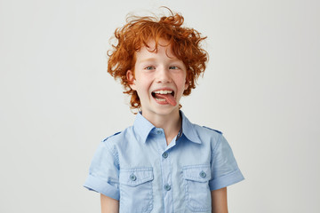 Portrait of silly little ginger boy in blue shirt with wild hair mowing eyes, smiling and showing tongue in camera,making funny faces.