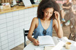 © Cookie Studio - Close up of young beautiful charming dark-skinned student girl with curly hair in stylish outfit sitting in cafe after long day at university, deinking coffe, doing her homework with sutisfied face