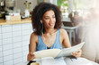 © Cookie Studio - Close up portrait young good-looking black-skinned woman with curly hair in casual clothes sitting in cafeteria, drinking coffee, listening music in earphones, looking through papers for work.